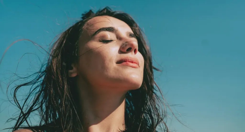 image: a woman standing in morning sunlight, their hair tousled by the wind.